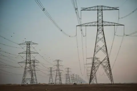 Linia wysokiego napięcia z wieżami energetycznymi, przypominającymi gigantyczne kogeneratory, ciągnie się przez pustynny krajobraz.