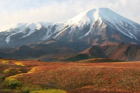 Majestätischer schneebedeckter Vulkan in einer weiten russischen Landschaft mit herbstlich gefärbten Hügeln im Vordergrund.