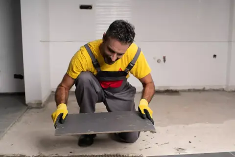 Obrero con guantes amarillos prepara un azulejo gris para pegar con Sikaflex en una obra.