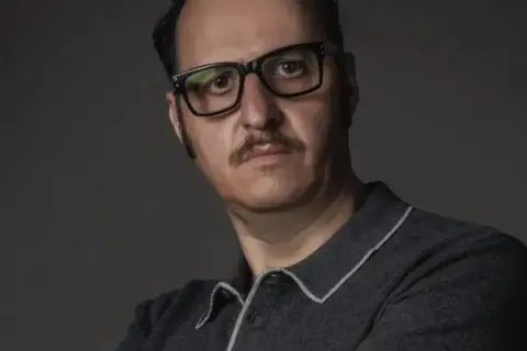 Ritratto di Fausto Russo Alesi con occhiali neri e baffi, indossa una polo grigia.