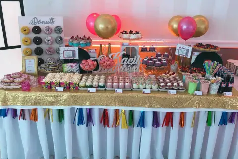 Słodki stół z donutami, babeczkami i deserami w słoiczkach, jak na wesele lata 80. Złoty obrus i kolorowe balony dodają blasku.