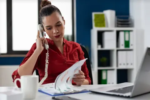 Kobieta w czerwonej bluzce prowadzi rozmowę telefoniczną, przeglądając dokumenty. Może to być element jej pracy związanej z twardą windykacją.