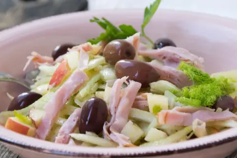 Frischer Fenchel Salat mit Apfelstücken, Schinkenstreifen und dunklen Oliven in einer rosa Schüssel.