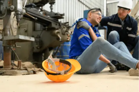 Robotnik po wypadku, kolega pomaga. Pomarańczowy kask leży obok. Sprawa dotyczy zadośćuczynienia a odszkodowania.