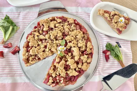 Ein köstlicher Rhabarberkuchen mit Streuseln, garniert mit kleinen Gänseblümchen. Frischer Rhabarber liegt daneben.