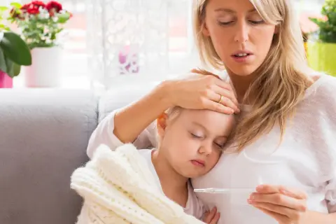 Zmartwiona mama sprawdza temperaturę chorej córki, która śpi otulona kocykiem. Myśli o podaniu Fervex dla dzieci.