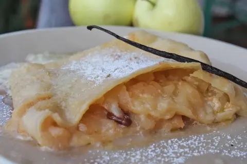 Ein Stück bayerischer Apfelstrudel, bestäubt mit Puderzucker und garniert mit einer Vanilleschote, daneben zwei Äpfel.