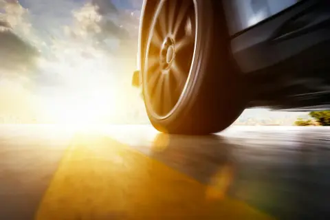 Roues d'une voiture en mouvement, le soleil rasant inonde la scène d'une lumière dorée, floutant le paysage urbain au loin.