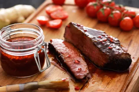 Saftigt kött med en glansig glaze, bredvid en burk med glaze recept. Tomater och ingefära i bakgrunden.