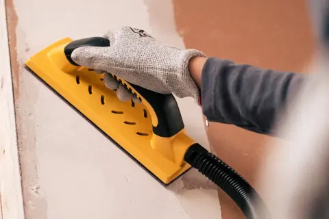Mano enguantada usando una lijadora amarilla para lijar paredes, preparando la superficie para pintar.