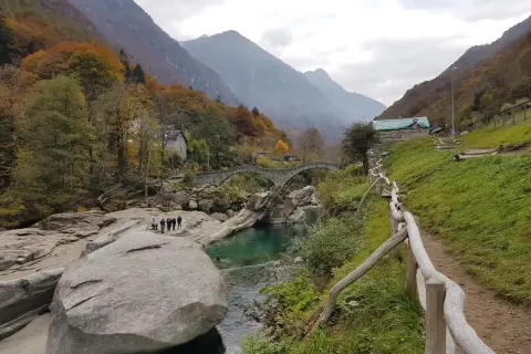 Sentiero sul monte Valdo, con un ponte ad arco in pietra, un fiume turchese e montagne sullo sfondo.