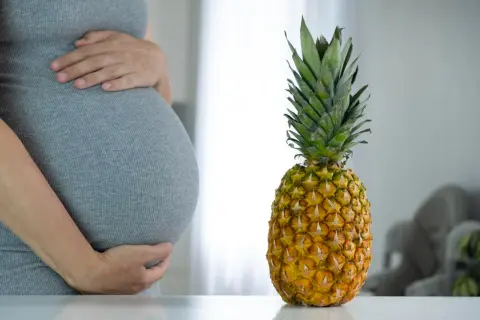 Ciężarna kobieta w szarej sukience trzyma się za brzuch obok ananasa.