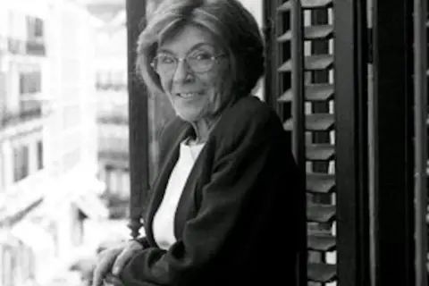 Carmen Rico Godoy, con gafas y chaqueta, mira a la cámara desde un balcón. Parece una escena de "Carmen Rico Godoy abandonada".