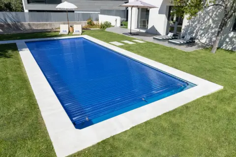 Piscina cubierta con persiana azul, lista para climatizar y disfrutar. Césped verde y tumbonas invitan al relax.