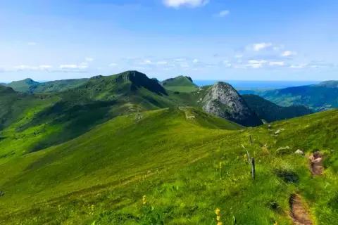 Paysage de montagnes verdoyantes sous un ciel bleu, idéal pour les plus belles randonnées dans le Cantal. Un sentier serpente sur la colline.