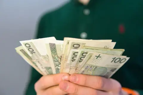 Dłonie trzymające plik banknotów 100 zł. Zastanawiasz się, ile wynosi renta rodzinna dla dziecka?
