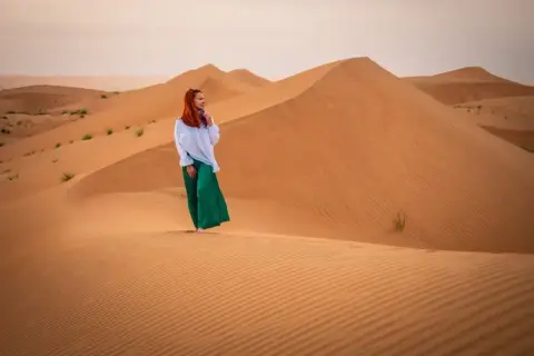 Kobieta w białej bluzce i zielonych spodniach stoi na piaszczystej wydmie. Czy Oman jest bezpieczny? Tak, to piękny kraj do zwiedzania.