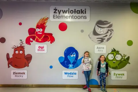 Dwie dziewczynki pozują na tle ściany z postaciami z bajki "Żywiołaki Elementoons". Widać Ogi Blaze, Ziemka Rocky, Wodzię Flow, Aero Airy i Żywka Zippy.