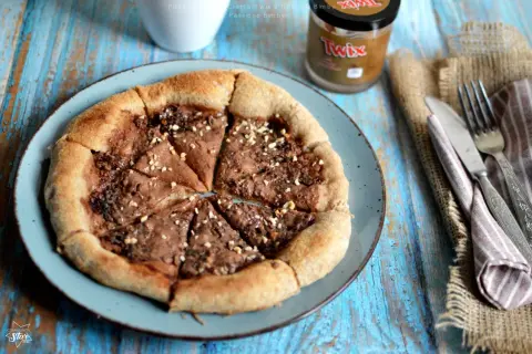 Pizza al cioccolato con crema Twix e nocciole, cosparsa di granella croccante. Un dolce goloso pronto da gustare.