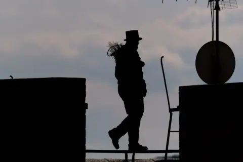 Silhouetten eines Bezirksschornsteinfegers mit Zylinder und Bürste auf dem Dach, der über ein Geländer geht.