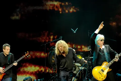Trzech muzyków na scenie podczas koncertu Led Zeppelin. John Paul Jones gra na basie, Robert Plant śpiewa, a Jimmy Page z uniesioną ręką gra na gitarze.
