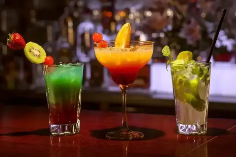 Trzy kolorowe drinki: zielono-czerwony z owocami, pomarańczowo-czerwony w kieliszku oraz zielony mojito.