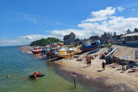 Słoneczny dzień nad polskim morzem. Kolorowe łodzie na plaży, tłumy ludzi, a na wodzie skuter wodny.