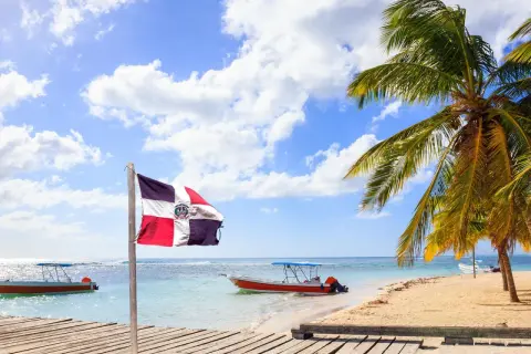 Dominikanische Flagge weht am Strand. Boote im türkisfarbenen Meer. Palmen. Hier ist es im Januar warm.