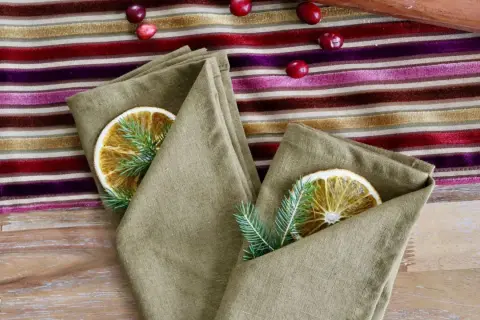 Un pliage serviette original avec une tranche d'orange séchée et une branche de sapin, sur une nappe rayée aux couleurs vives.
