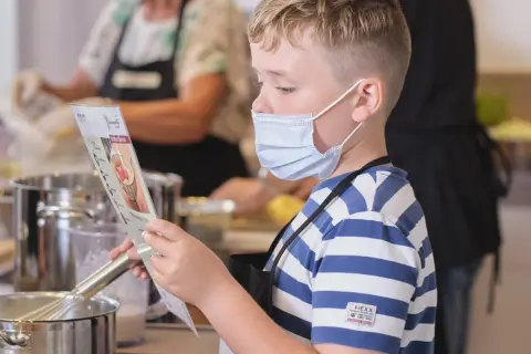 Junge mit Maske liest Rezept, während andere kinder kochen selbst. Küchenutensilien und Töpfe sind zu sehen.