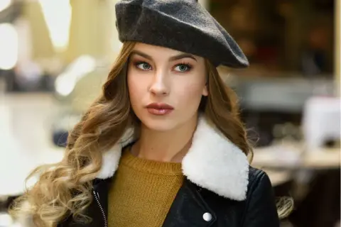 Elegancka kobieta w grafitowym beretu, z burzą blond loków i w czarnej ramonesce. Dowiedz się, jak nosić beret damski, by wyglądać stylowo.