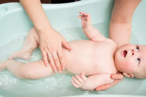 Rączki mamy delikatnie trzymają dziecko w wodzie w wanience dla niemowlaka. Maluch z zaciekawieniem patrzy w górę.