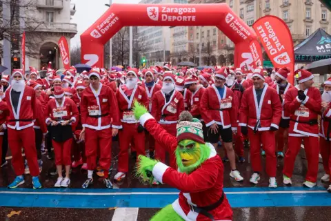 Multitud de personas disfrazadas de Papá Noel en Zaragoza, listas para una carrera. Un Grinch verde se une a la fiesta.