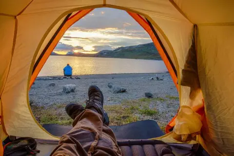 Blick aus dem Zelt auf einen See bei Sonnenuntergang. Eine Person sitzt am Ufer. Ja, darf man in Schweden wildcampen!