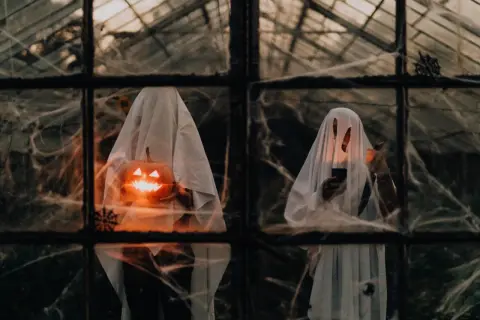 Zwei Geister mit Kürbissen und Kerzen, die durch ein Spinnennetz-verhangenes Fenster blicken. Ein Blick in die Ursprünge von Halloween.