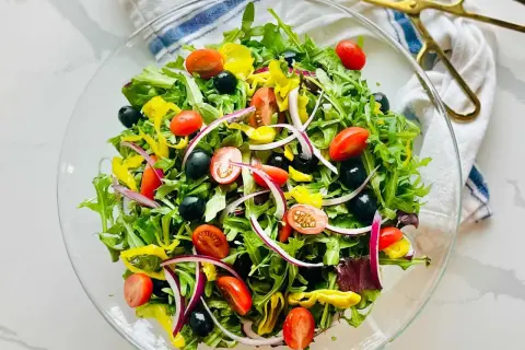 En fräsch italiensk sallad i en glasskål, fylld med gröna blad, körsbärstomater, svarta oliver, rödlök och gul paprika.