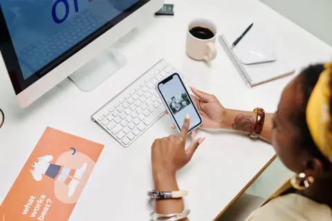 Une développeuse consulte son blog sur son téléphone, entourée d'un ordinateur, d'une tasse de café et de documents de conception.