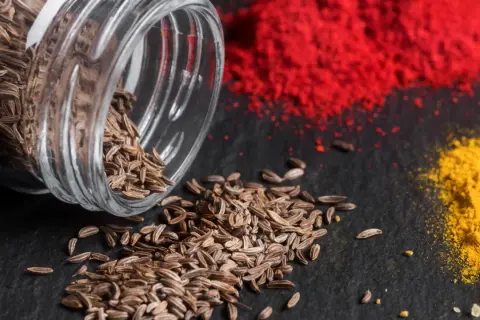 Braune Kümmelsamen quellen aus einem Glas, daneben leuchtend rote und gelbe Gewürzpulver. Was ist Cumin? Es ist dieses aromatische Gewürz!
