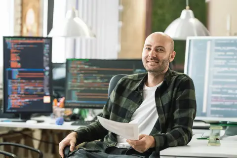 Ein Mann mit Glatze, der ein Dokument hält, sitzt vor mehreren Computermonitoren mit Code. Er könnte einen Techniker im Fernstudium bei der IHK absolvieren.