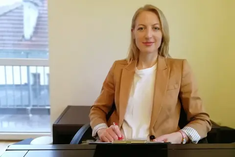Kobieta w beżowej marynarce siedzi przy biurku i pisze.