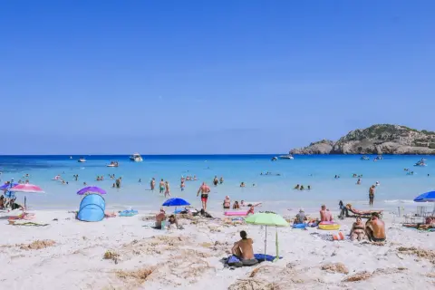 Słoneczna plaża Cala Agulla tętni życiem. Ludzie kąpią się w krystalicznie czystej wodzie, opalają na piasku i odpoczywają pod parasolami.