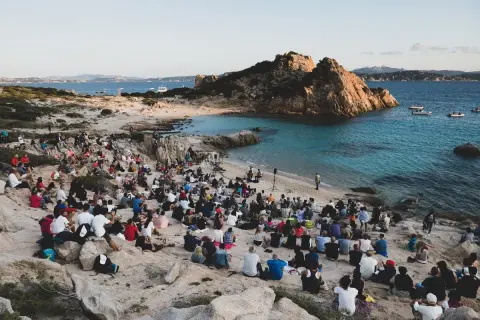Concerto sulla spiaggia in Sardegna, con un pubblico seduto tra le rocce che ascolta un cantante sardo.