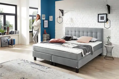 Modernes Boxspringbett in Grau, ideal für erholsamen Schlaf bei Bandscheibenvorfall. Eine Frau lehnt lässig daneben.