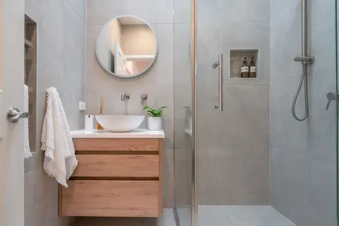 Bagno piccolo con lavabo a conca, mobile in legno, specchio rotondo e doccia con piastrelle grigie.