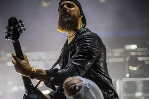 Charyzmatyczny gitarzysta z brodą podczas koncertu Bullet for My Valentine, w skórzanej kurtce i podartych spodniach, gra na gitarze.