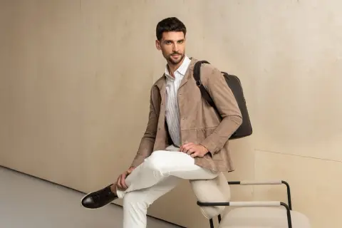 Elegancka stylizacja męska: beżowa kurtka z zamszu, białe spodnie, koszula w paski i czarny plecak.