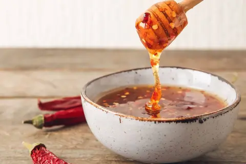 En träsked droppar gyllene chili honung ner i en skål. Torkade chilifrukter ligger bredvid.
