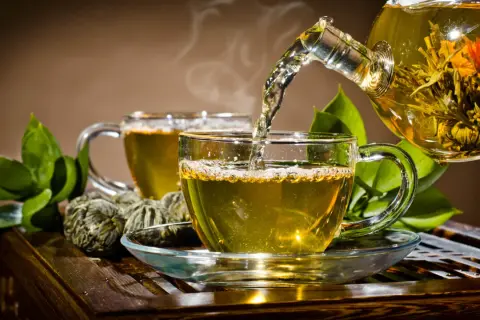 Gorąca herbata parzy się w szklanej filiżance, a z dzbanka wlewa się aromatyczny napar. Obok leżą zielone kulki herbaty.