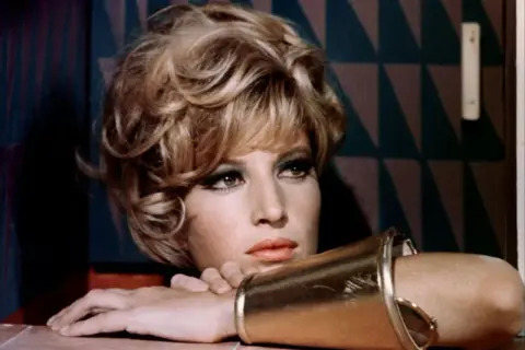 Monica Vitti, con un bracciale dorato e un'espressione pensierosa, sfoggia un look audace con capelli biondi cotonati e trucco marcato.