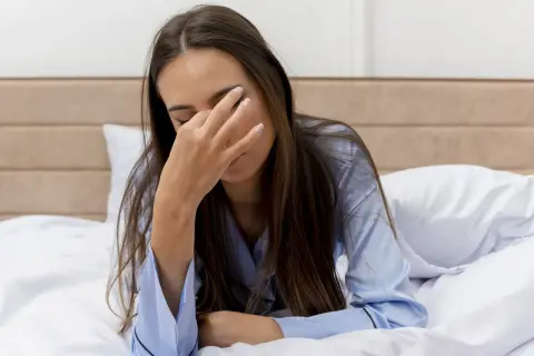 Een vrouw ligt moe in bed, haar hand voor haar ogen. Ze draagt een blauwe pyjama.
